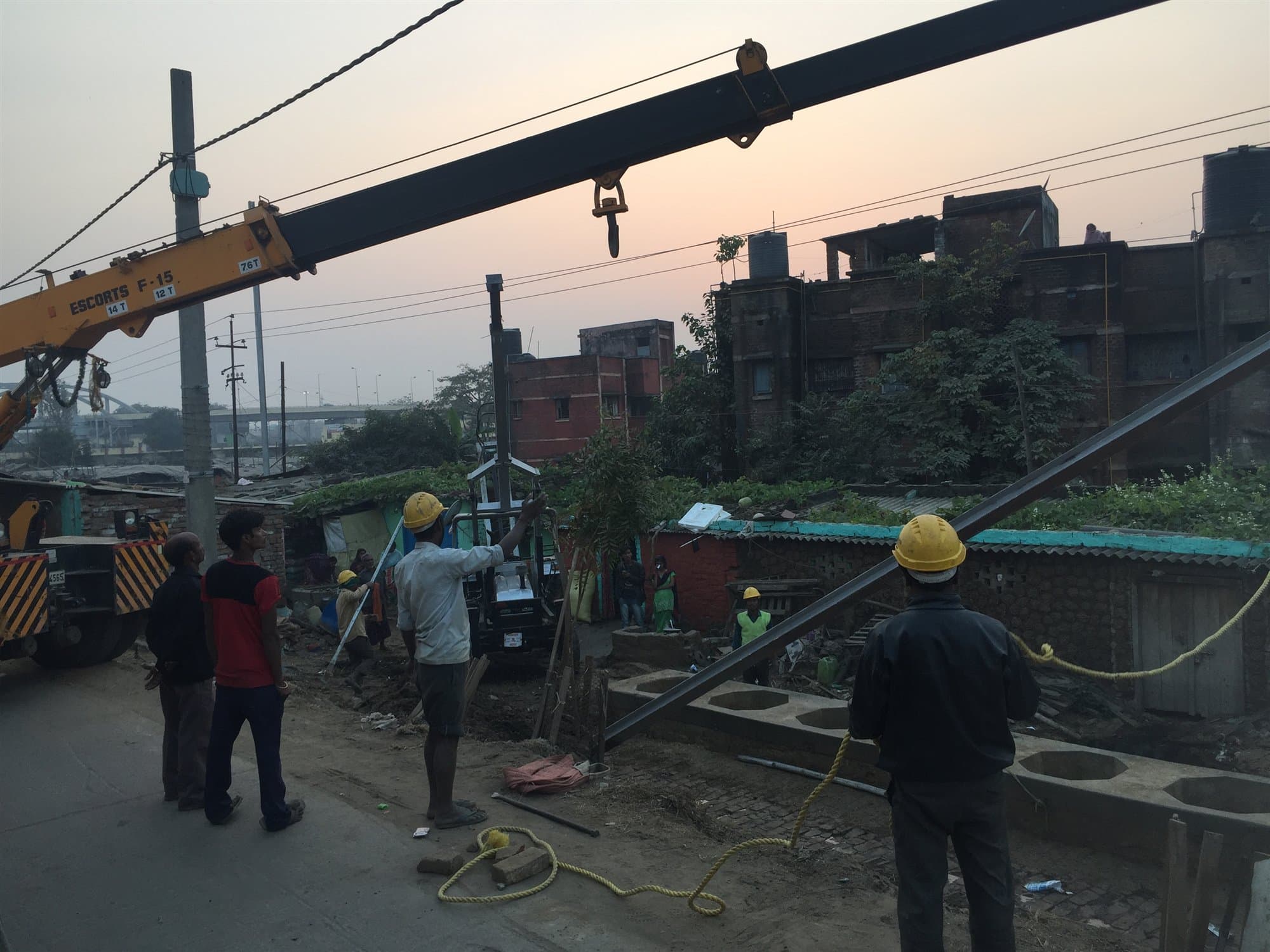 A hydraulic truck crane holding a long steel girder above a street at dusk, with a crew watching from below