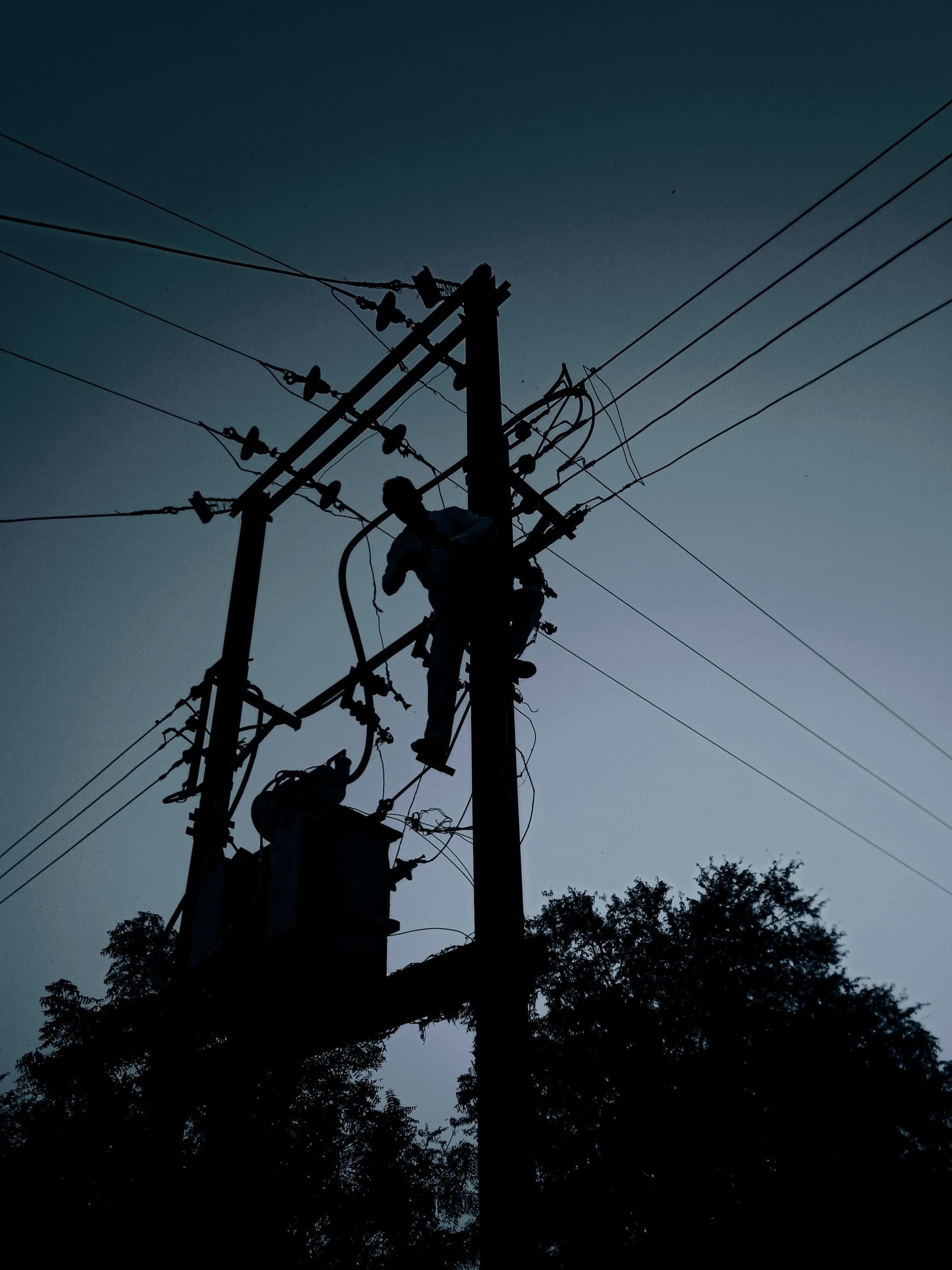 A lineman working at the top of a distribution pole at dusk, silhouetted against the sky